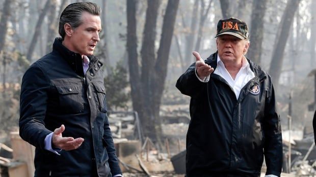 A cleanshaven older man wearing a baseball cap that says USA gestures with his hand while walking outside with a younger, clean-shaven man. They are in an area with large trees in the background.