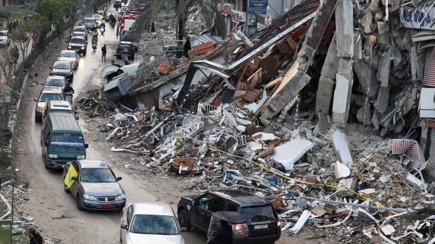 A line of cars drives along an urban street filled with destroyed buildings. 