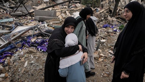 A woman in black hugs a child in front of debris of a damaged building. A few other women are nearby.