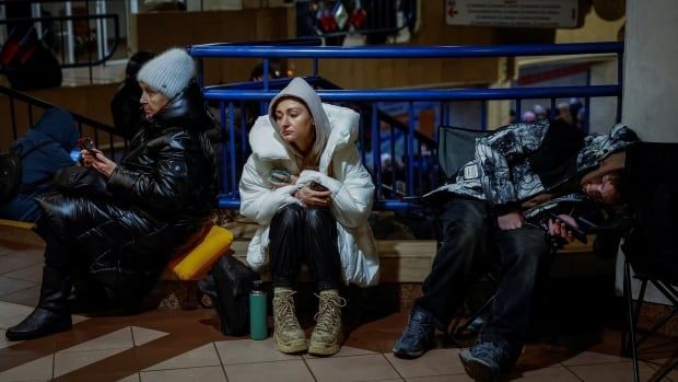 Three women in winter coats sit near a railing in a metro station.