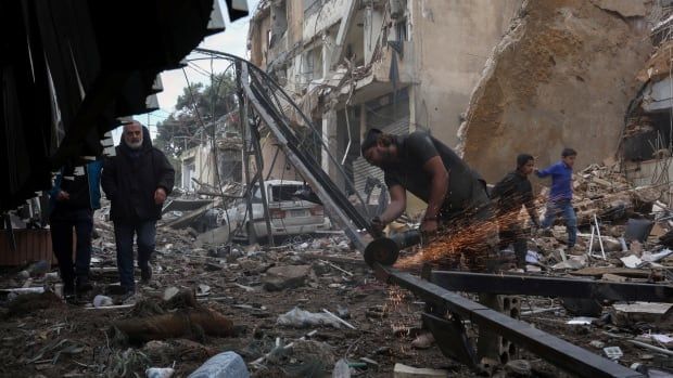 People walk on the rubble of damaged buildings as a man cuts steel.