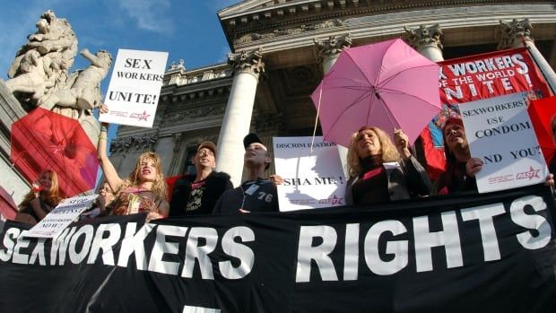 A group of people stand on the steps of an ornate white building holding signs, umbrellas and a large banner that reads: "SEX WORKERS RIGHTS"