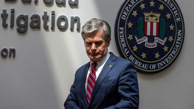 A man in a blue suit stands in the centre of the image in front of a wall that says "Bureau" and has a large seal image of the FBI on it. A beam of light on the wall illuminates him slightly dramatically as he looks down. 