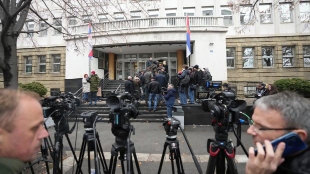 Two men are seen in the foreground, one holding a cellphone to his ear, near a bunch of standup tripods in front of a building. In the backgrounds, a few dozen people are shown on the steps of a building.