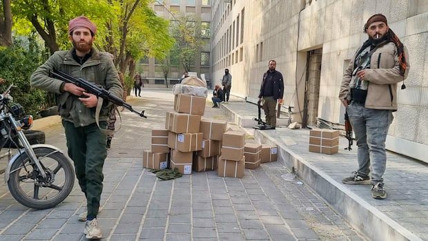 Several men with long guns stand in a street, guarding a pile of boxes.