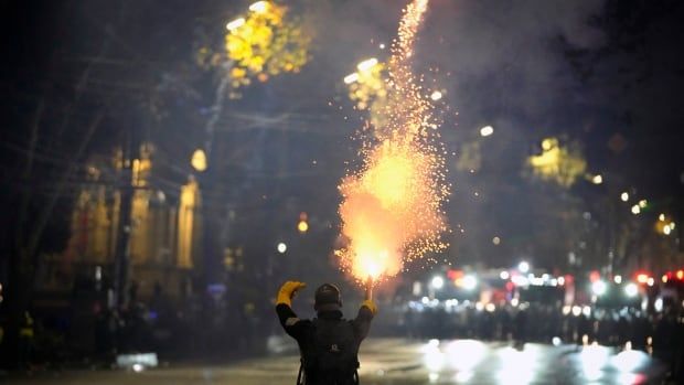 A person wearing dark clothing and bright yellow gloves stands with their back to the camera holding a firework and facing off against a line of law enforcement with shields in the distance. 