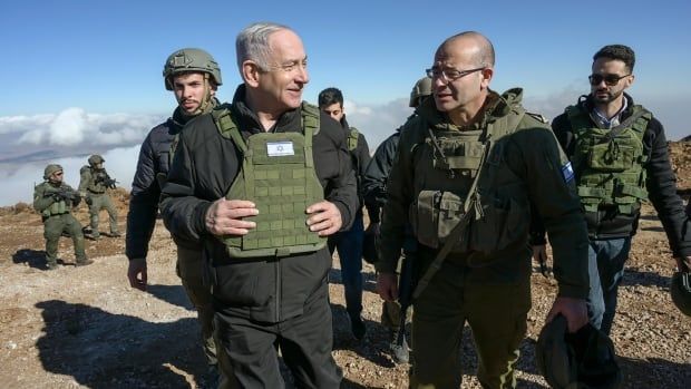 Two men wearing green military vests walk on a rocky landscape, flanked by soldiers.