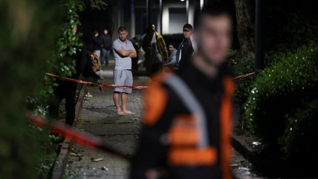 A person in the background crosses their arm while standing behind police tape as an official works in the foreground.