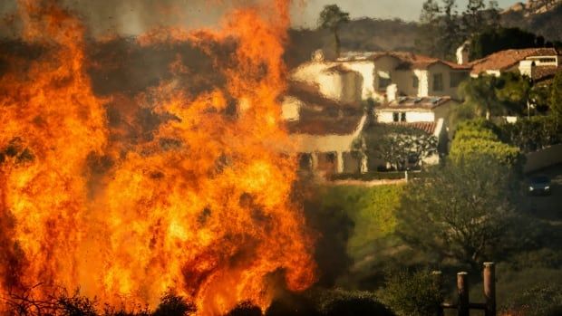 In the foreground is a lot of flames, mostly at the left side of the image. In the distance, warped by the heat of the flames, a large house or multiple houses are visible. 