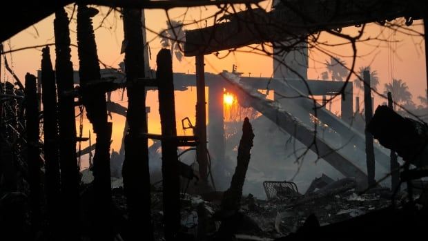 The sun is seen in the distance through the silhouette of black, burned support beams in what is left of a structure. Grey dust or smoke rises above the rubble. 