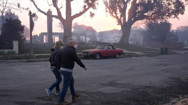 two people walk on a road, both wearing medical masks over their noses and mouths. there is a car parked on the street in front of burned buildings