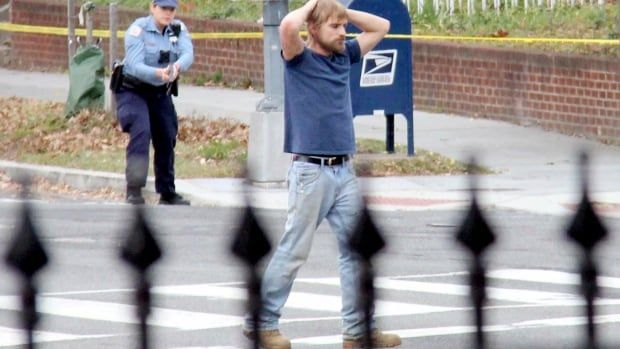 A bearded Caucasian man in a tshirt and jeans places his arms over his head on a city street as a police officer is shown in the background with a weapon drawn.