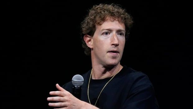 A curly-haired, clean shaven man is shown sitting and gesturing with his left hand while holding a microphone with his right hand.