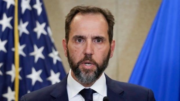 A bearded man wearing a suit and tie looks into the camera while speaking on a stage.