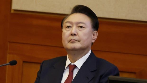 An older cleanshaven Asian man wearing a suit and tie is shown seated inside a room.