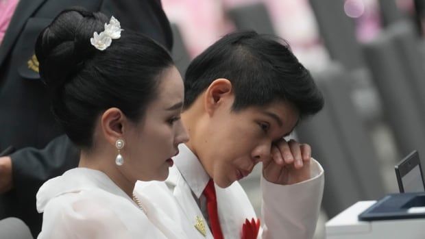 A couple side by side in a large room. The woman on the right wipes tears from her eyes. 