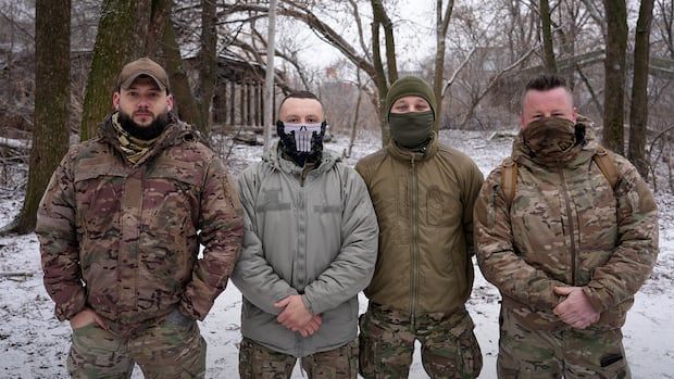 Four men in military fatigues stand in a snowy field. Two have their faces partially covered.