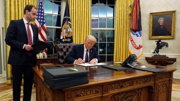 A man seated at a large desk signs a piece of paper while another man stands to his right. 