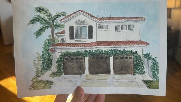 A hand holds up a 5-by-7 watercolour painting of a two-storey, California-style house,  covered in ivy, with three garage doors, and palm tree next to it. 