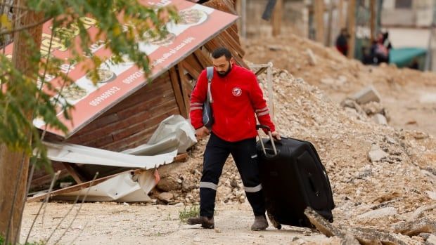 A man in a red sweatshirt walks with a suitcase on a damaged street. He also has a backpack slung over his shoulder.