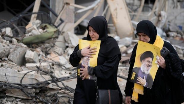 Two women hold portraits of a Hezbollah leader as they walk through a destroyed neighbourhood.