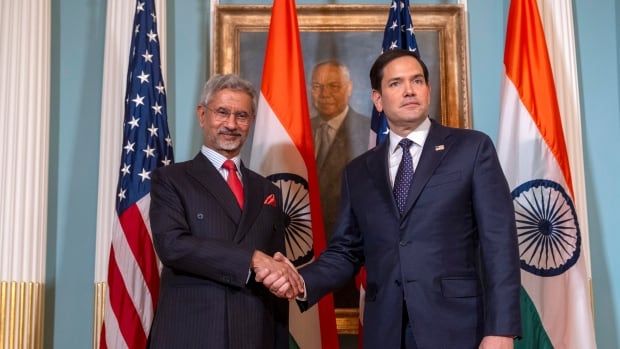 A bearded man in glasses shakes the hand of a dark haired cleanshaven man. Both are wearing suit and tie, in front of India and U.S. flags and a framed portrait of a bespectacled man.