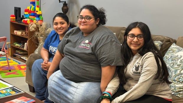 A woman sits on a couch, with her two teenage daughters sitting either side of her.