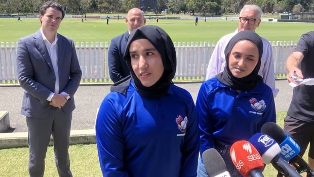 Two women in blue uniforms speak in front of microphones while three men stand behind them. A cricket pitch is shown in the background.