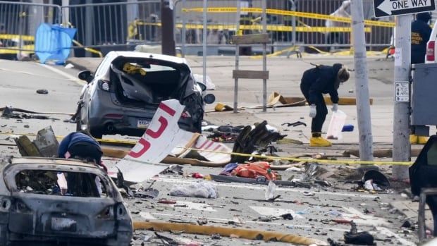 A person collects aircraft wreckage on the street near damaged cars.