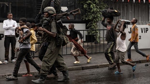 A helmeted man with a large gun strapped on his fatigues is shown walking down the street surrounded by other dark complected people, civilians which include children.