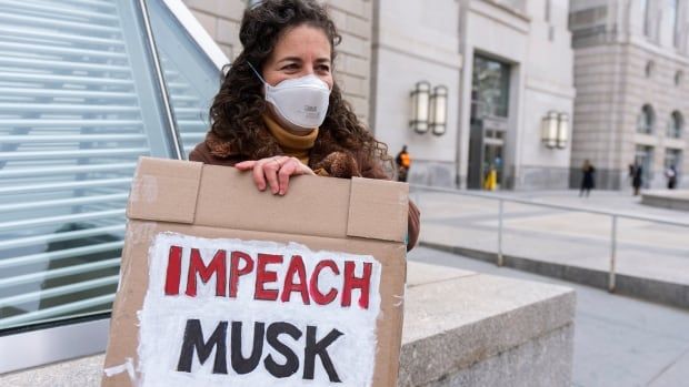 A woman with shoulder length brown hair wearing a mask over her face stands outside a building and holds up up a sign that says 'Impeach Musk.'