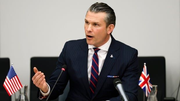 A cleanshaven, dark haired man in a suit and tie gestures while speaking, seated at a table between small American and British flags.