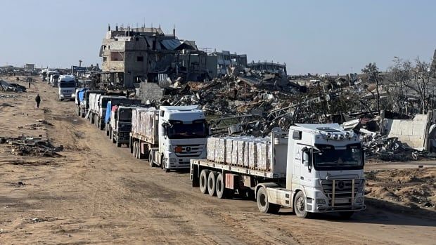 Trucks are shown travelling on a dirt rood amid heavily damaged buildings and concrete debris.