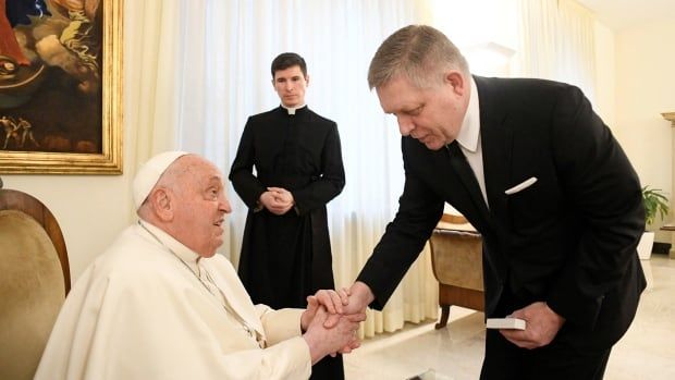 The pope, seated and dressed in white garments, clasps the right hand of a cleanshaven older man in a suit and tie who is standing. Nearby a priest in dark religious outfit stands.