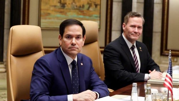 Two cleanshaven men wearing suit and tie are shown seated at a table, with a small American flag in front of them.