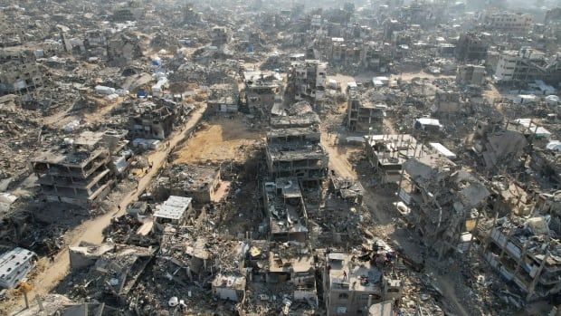 An aerial shot shows countless grey, destroyed buildings, most of them either skeletons or half falling down. Others are completely smashed into piles of rubble. 