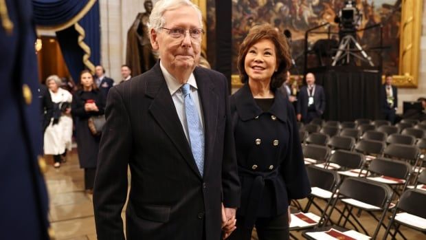 An older white haired man in glasses wearing a suit and tie walks next to an Asian woman wearing a blazer.