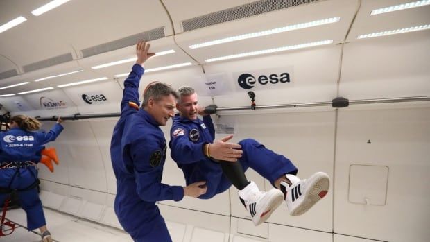 Two men experiencing weightlessness on a parabolic flight. They wear blue jumpsuits, and one has a prosthetic leg.