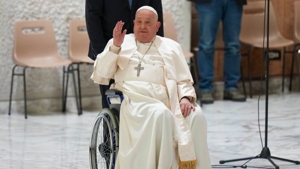 A person in religious garb waves while being pushed in a wheelchair.