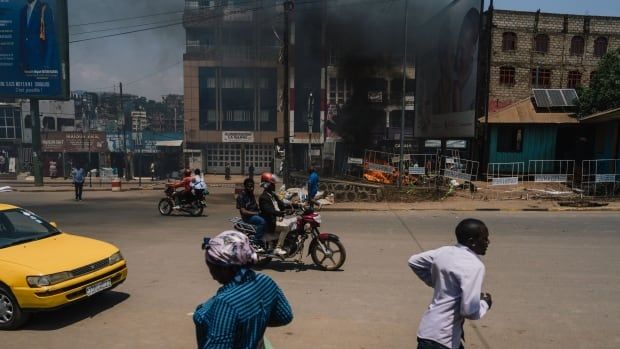 Several people are shown on a city street, some appear to be running, as in the background a blackened and charred lowrise building is shown.