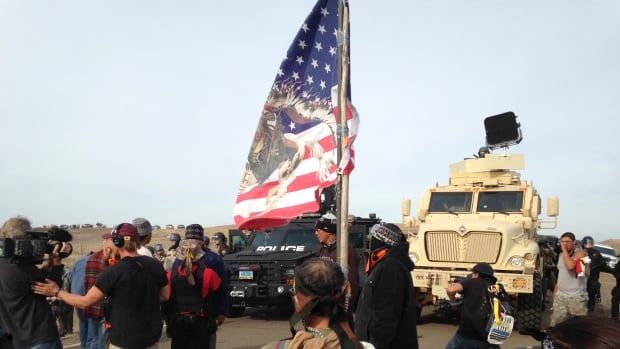 Several people are shown standing outside in a rural area in front of vehicles. One hoists a large flag that is blue and red with an eagle on it.