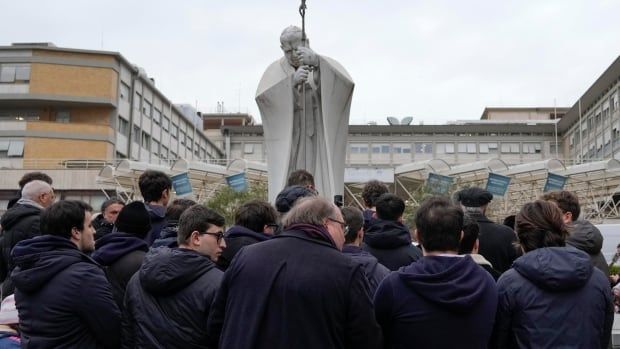 People in black clothes pray by a white statue of Pope John Paul II.