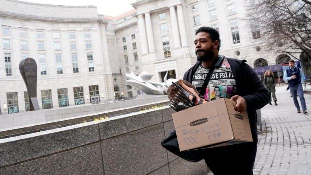 A bearded man carries a box containing items outside a building.