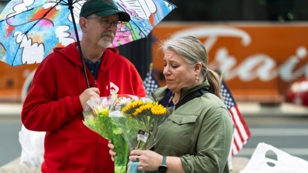 A woman in a coat looks forlorn while holding a yellow bouquet of flowes. Behind her is a man and some American flags.