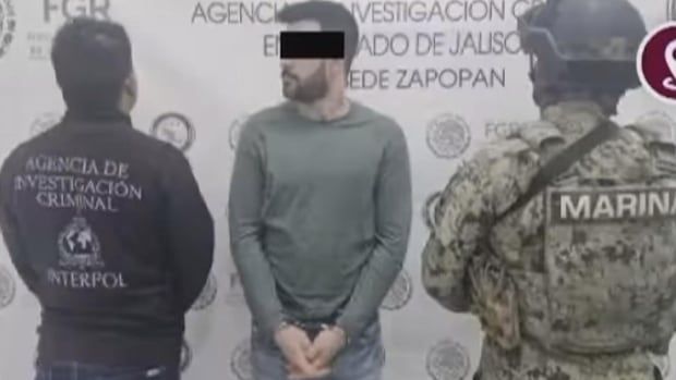 A man in handcuffs is surrounded by someone in an INTERPOL jacket and a soldier in camouflage