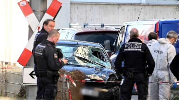 Several people, including a few in police uniforms, stand around a car that has a smashed windshield.