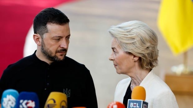 A bearded, dark haired man wearing dark clothing and an older, blonde haired woman turn to face each other in close proximity near a bank of microphones.