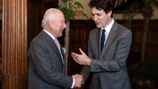 Two people shake hands in a formal room with a wooden pillar to the side and large tree-like plants in the background.