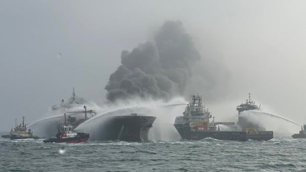 A number of boats are gathered around the blackened, smoking prow of a large ship at sea, and are spraying jets of water at it. 