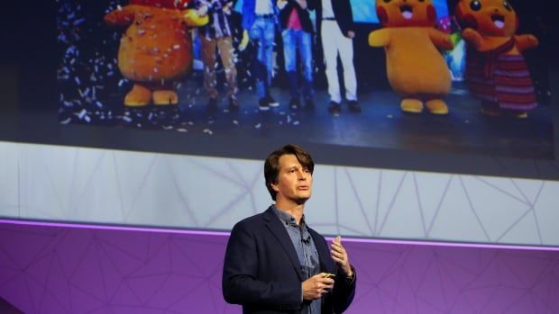 A man with short brown hair weraing a blue blazer and shirt stands on a stage under a giant projection of Pokemon characters.    